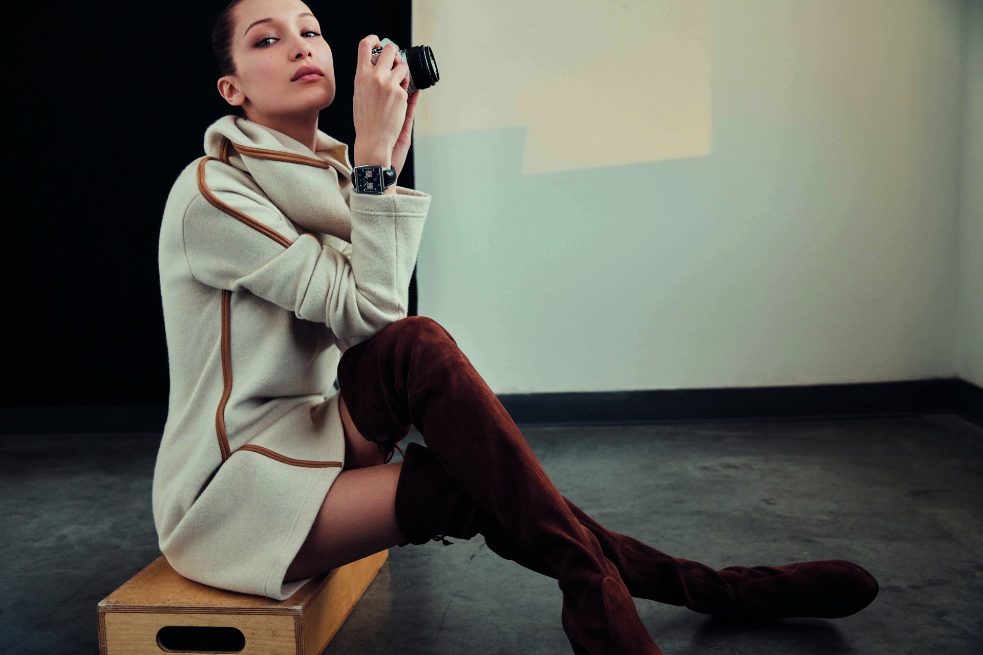 HD desktop wallpaper of celebrity Bella Hadid seated on a box wearing thigh-high boots and a wristwatch, holding a camera against a minimalist background.