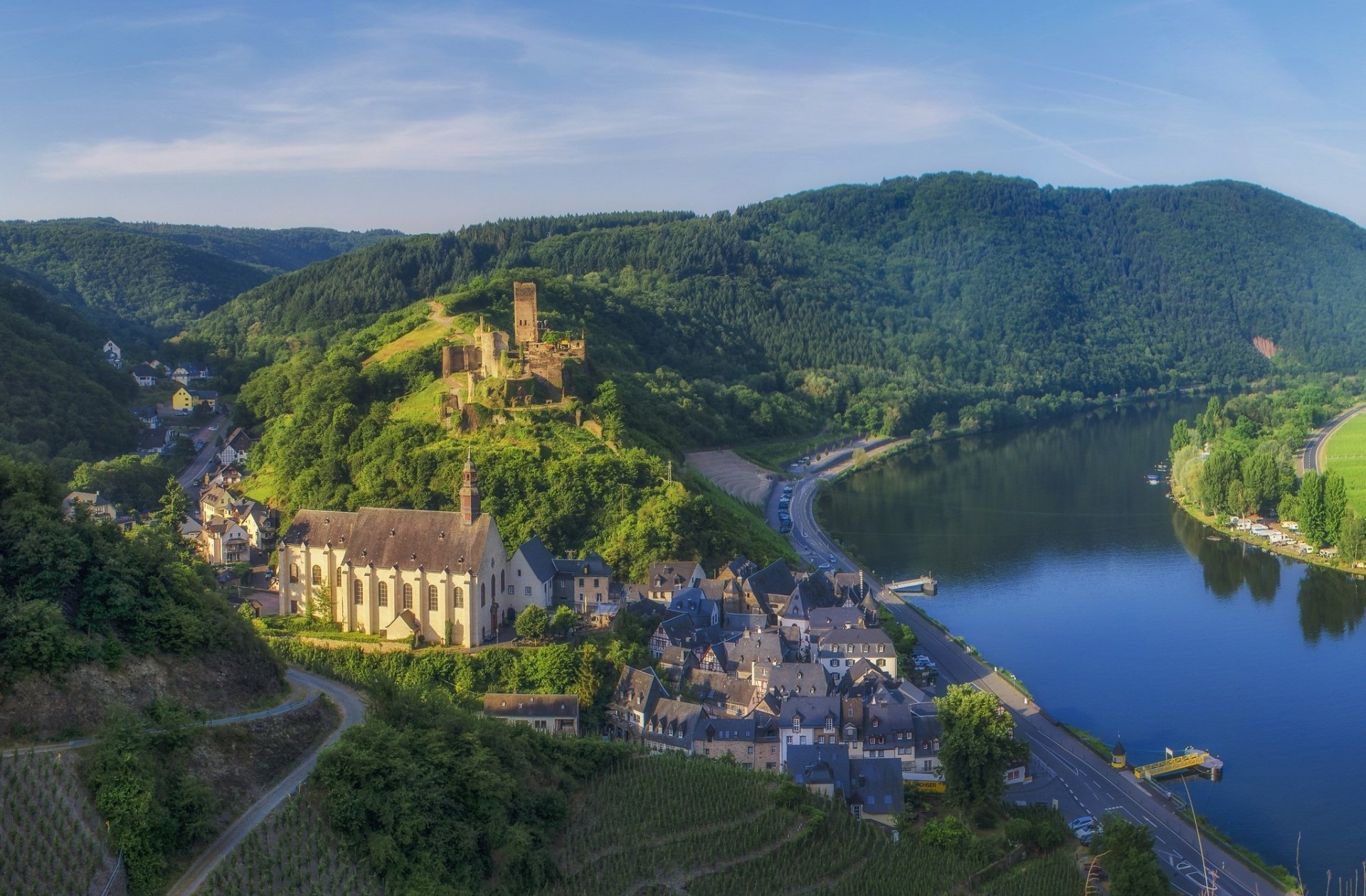 HD PC desktop wallpaper featuring a man-made village nestled by a river, surrounded by lush green hills under a clear blue sky.