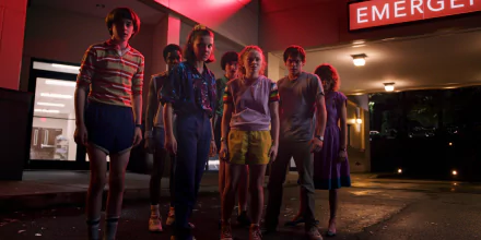 HD desktop wallpaper featuring characters from the TV show Stranger Things standing together outside a brightly lit building with an 'Emergency' sign.