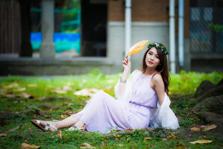 Asian woman with long brunette hair, wearing a lavender dress and floral wreath, sits on the grass, holding a handheld fan. The serene setting makes it a captivating HD desktop wallpaper background.