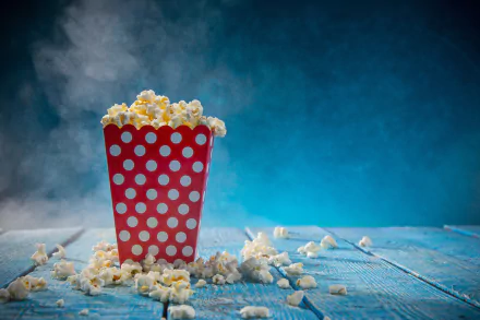 5K Ultra HD PC desktop wallpaper: a red polka-dot tub of popcorn spilling across a blue wooden table, scattered kernels and wisps of steam against a teal background.