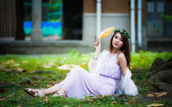 Asian woman with long brunette hair, wearing a lavender dress and floral wreath, sits on the grass, holding a handheld fan. The serene setting makes it a captivating HD desktop wallpaper background.