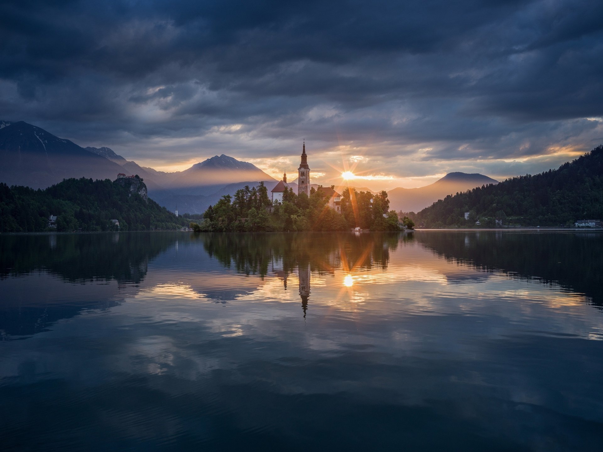 Sunlight shines over the Assumption of Mary Church on a small island in Slovenia, reflecting on the calm water beneath a dramatic sky.