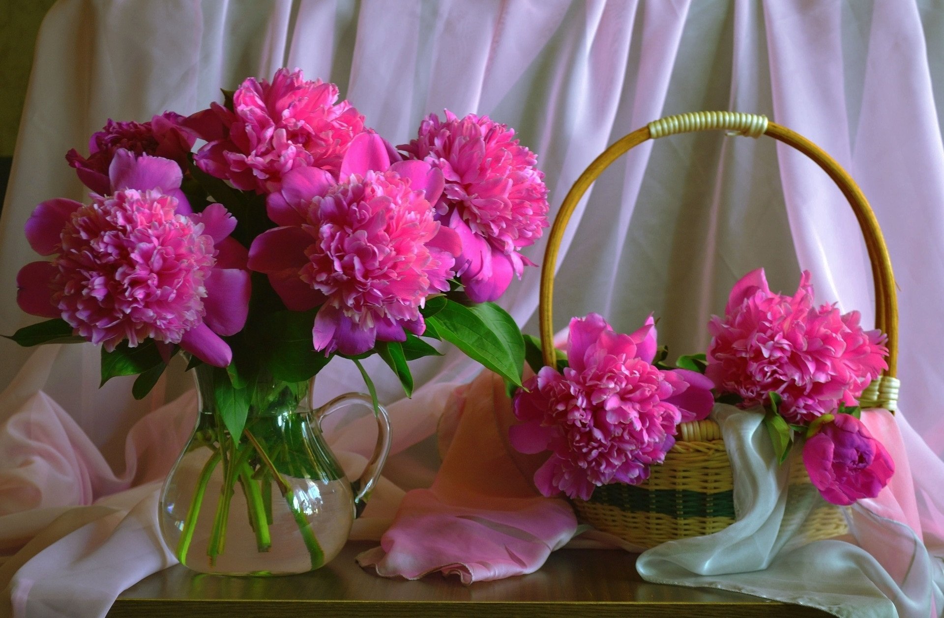 HD desktop wallpaper featuring a still life of vibrant pink flowers arranged in a glass pitcher and a woven basket, set against soft draped fabric.
