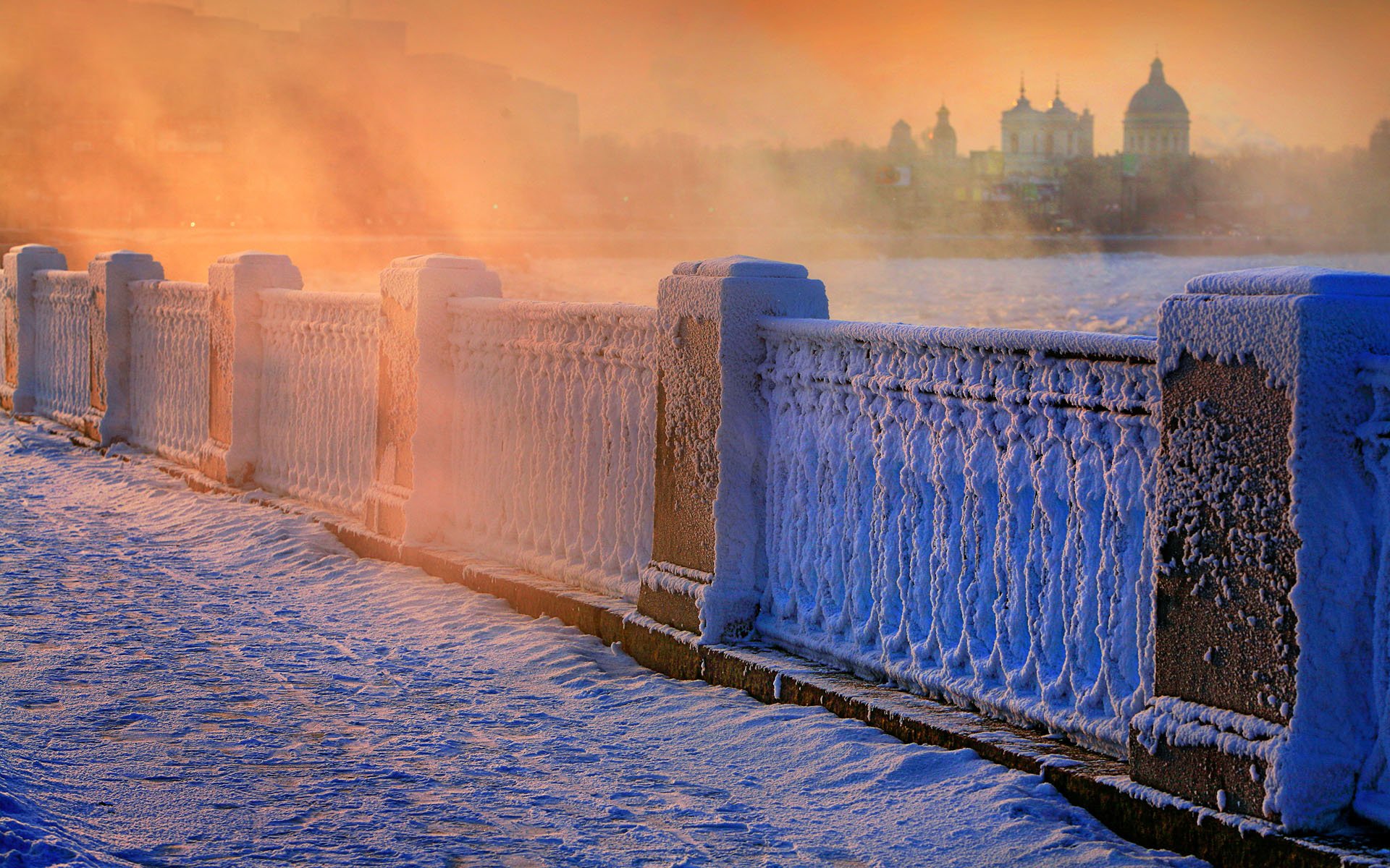 HD desktop wallpaper of a winter scene with a snow-covered ornate fence and misty city skyline bathed in warm orange light.