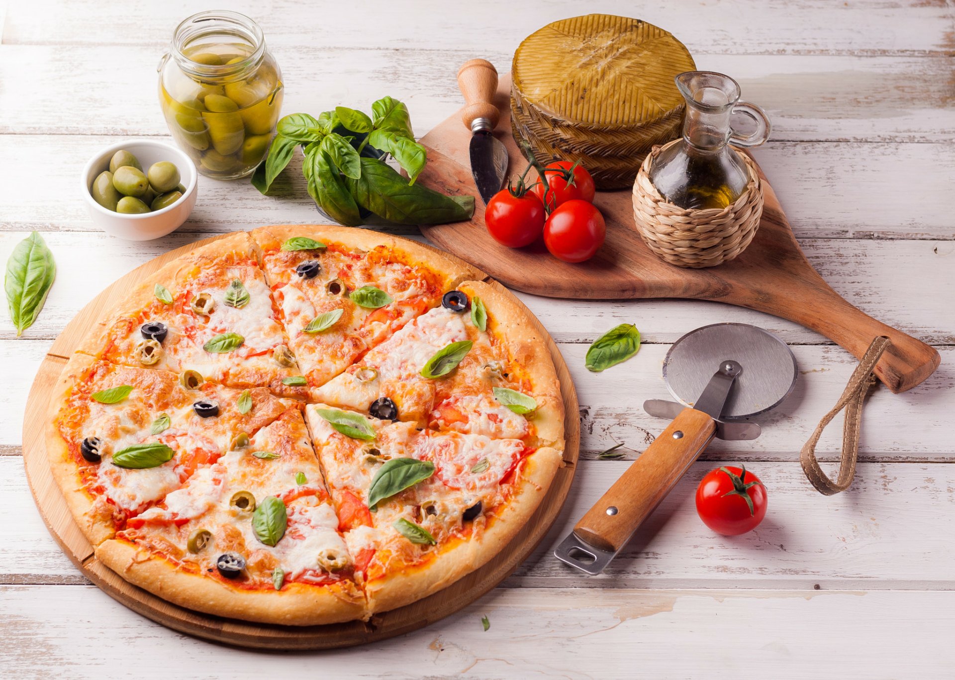 HD still life desktop wallpaper featuring a freshly baked pizza topped with olives, tomatoes, and basil, surrounded by olive oil, whole olives, and fresh ingredients.