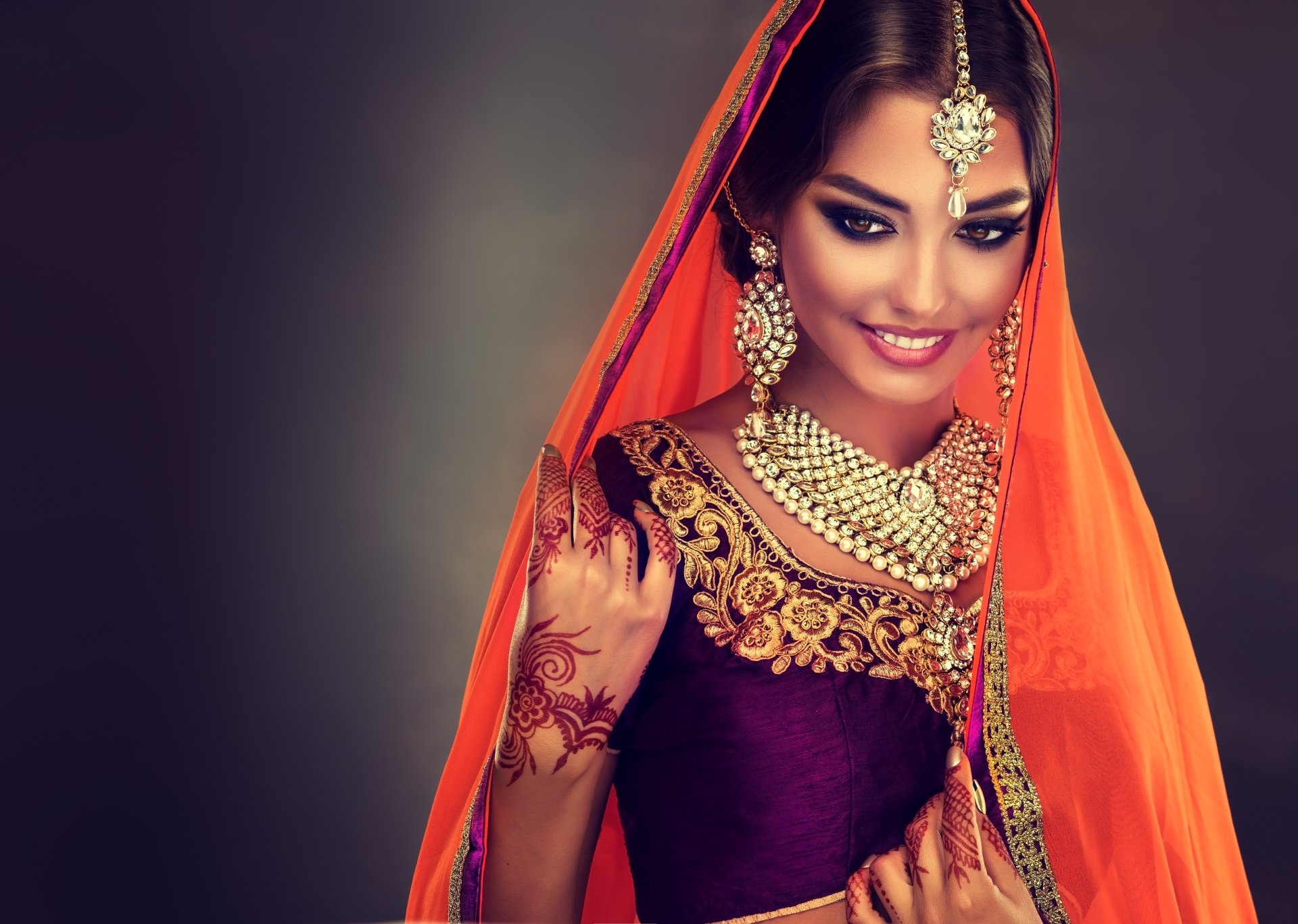 4K Ultra HD PC desktop wallpaper of a smiling Indian model in a saree — black hair, brown eyes, ornate necklace and earrings, mehndi on her hand and a vibrant orange veil.