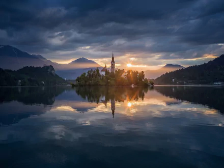 Sunlight shines over the Assumption of Mary Church on a small island in Slovenia, reflecting on the calm water beneath a dramatic sky.