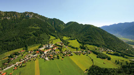 Aerial HD desktop wallpaper of a man-made Bavarian village nestled in the German Alps, with green fields, scattered houses and forested hills.