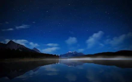 A serene nighttime landscape featuring a starry sky, wispy clouds, and a tranquil lake reflecting mountains and fog, capturing the beauty of nature in HD clarity.