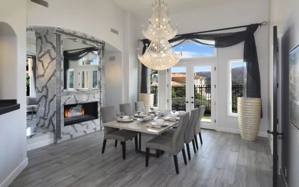 Modern dining and living room featuring a crystal chandelier, gray upholstered chairs, a rectangular dining table, marble fireplace, and large windows, captured in 4K Ultra HD.