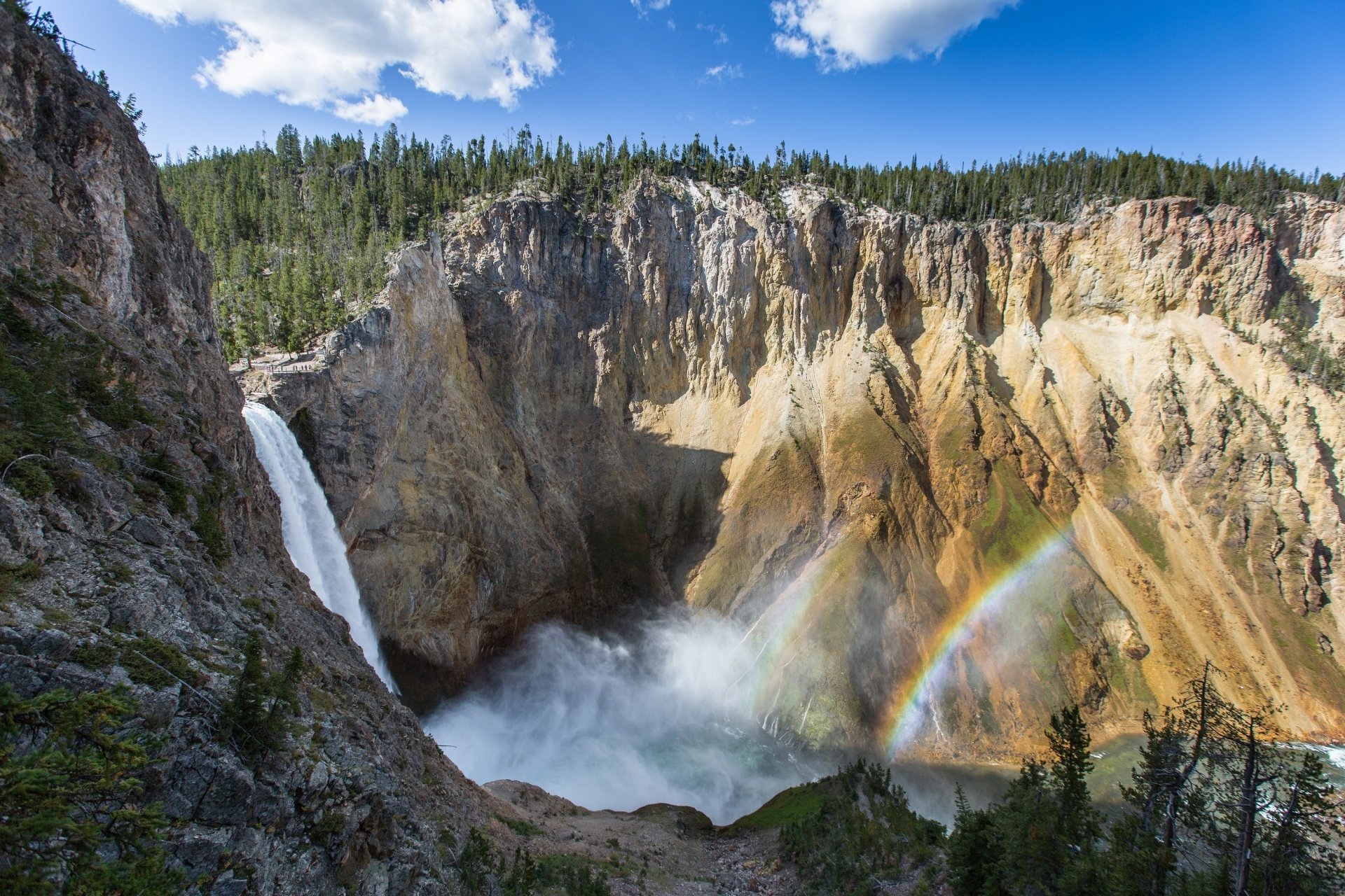 HD desktop wallpaper of a stunning waterfall cascading into a rocky canyon with a vibrant rainbow under a bright blue sky in a USA national park setting.