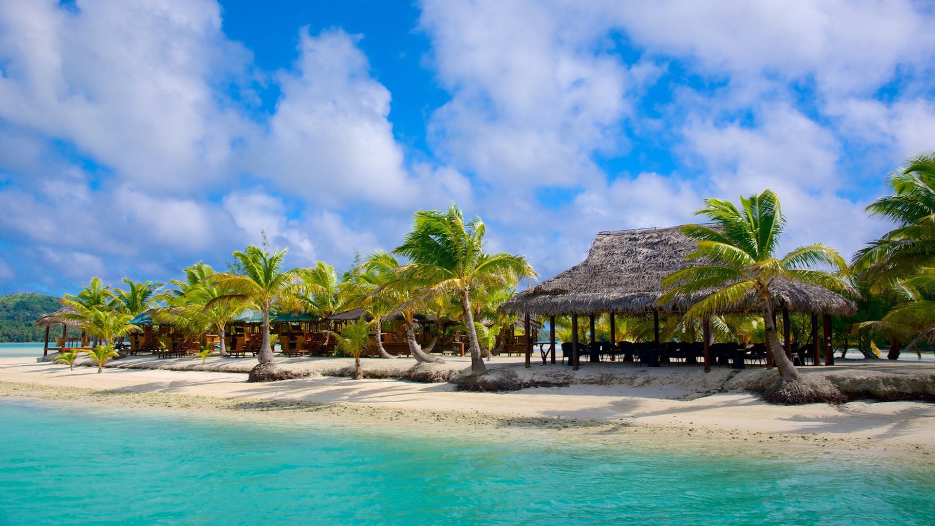 HD desktop wallpaper showing a tropical man-made resort with palm trees, sandy beach, and clear turquoise water under a bright blue sky with scattered clouds.