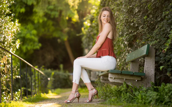 A blonde woman wearing high heels and white pants sits on a bench in a sunlit outdoor setting with lush greenery, captured in 4K Ultra HD with depth of field.