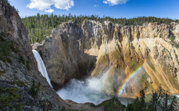 HD desktop wallpaper of a stunning waterfall cascading into a rocky canyon with a vibrant rainbow under a bright blue sky in a USA national park setting.