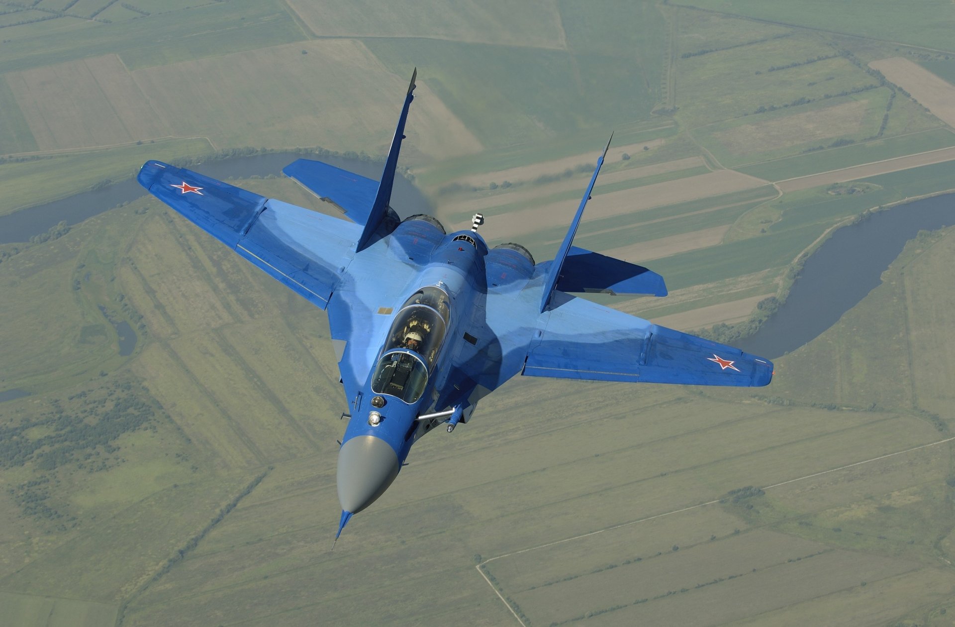HD desktop wallpaper featuring a blue Mikoyan MiG-29 military jet fighter warplane flying over a patchwork of fields.