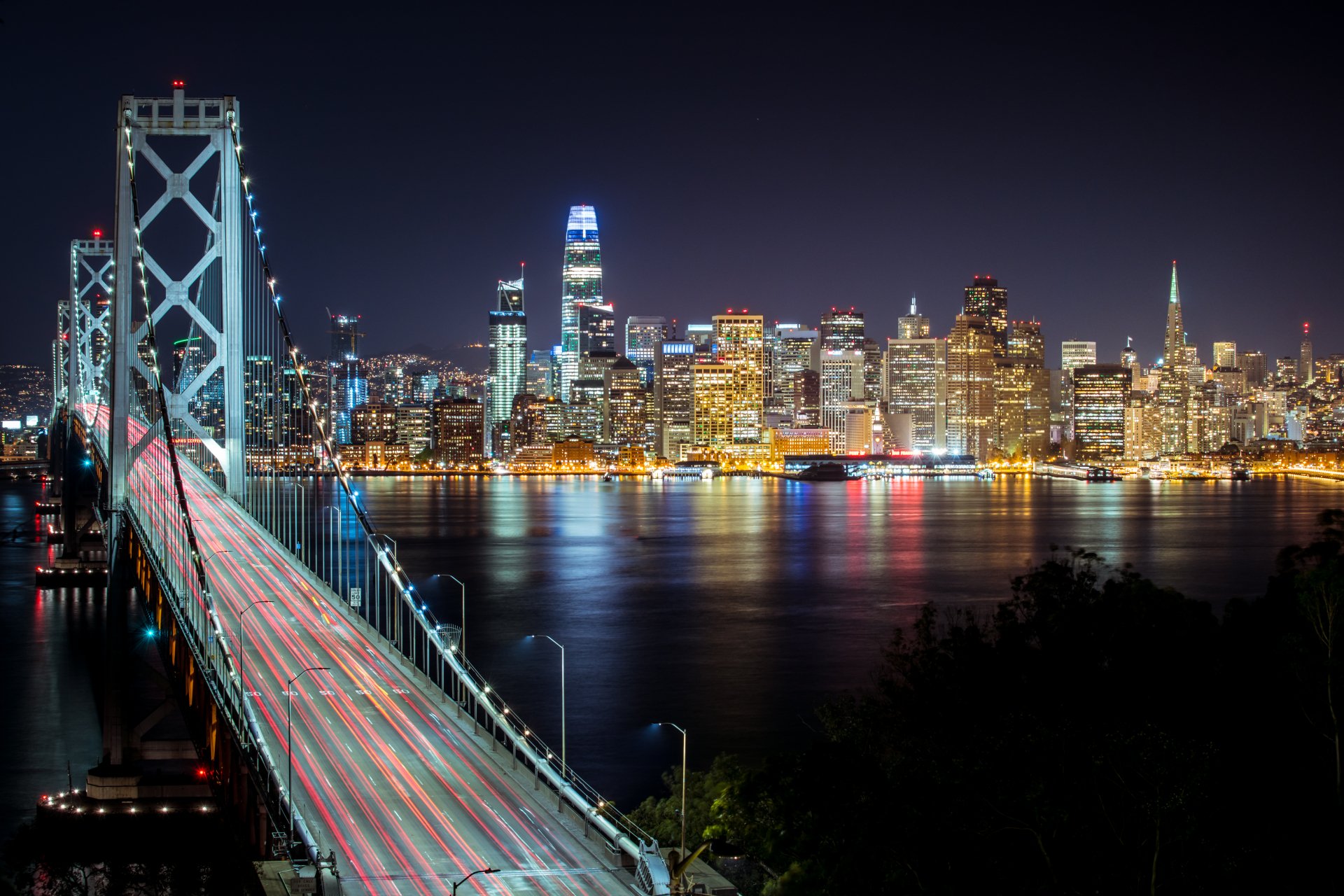 8K Ultra HD PC desktop wallpaper of a man-made cityscape: illuminated Bay Bridge stretching toward a glittering night skyline reflected on the bay.