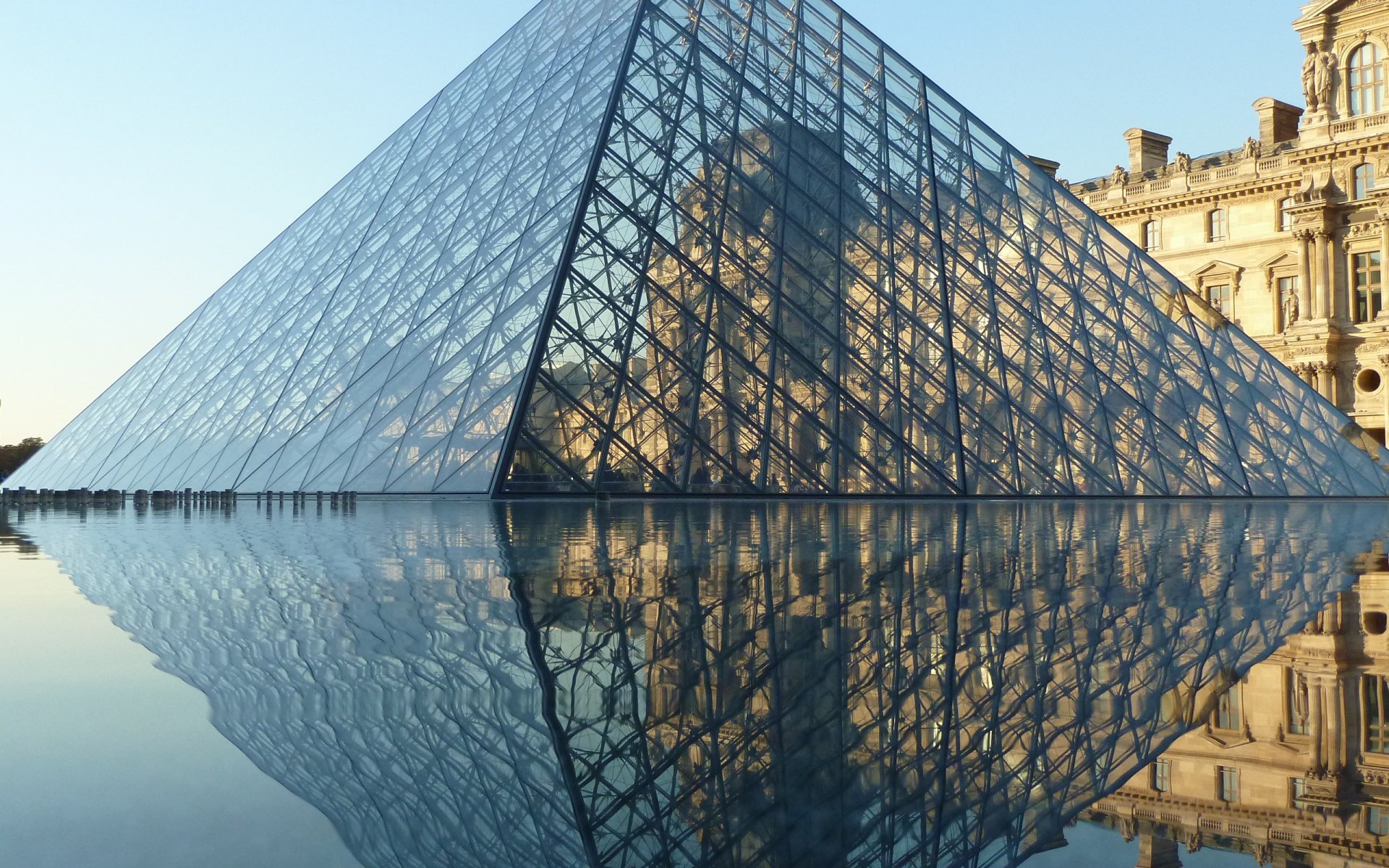 HD wallpaper of the Louvre Pyramid in Paris, France, showcasing its glass structure reflected in the fountain water beside the historic museum building.