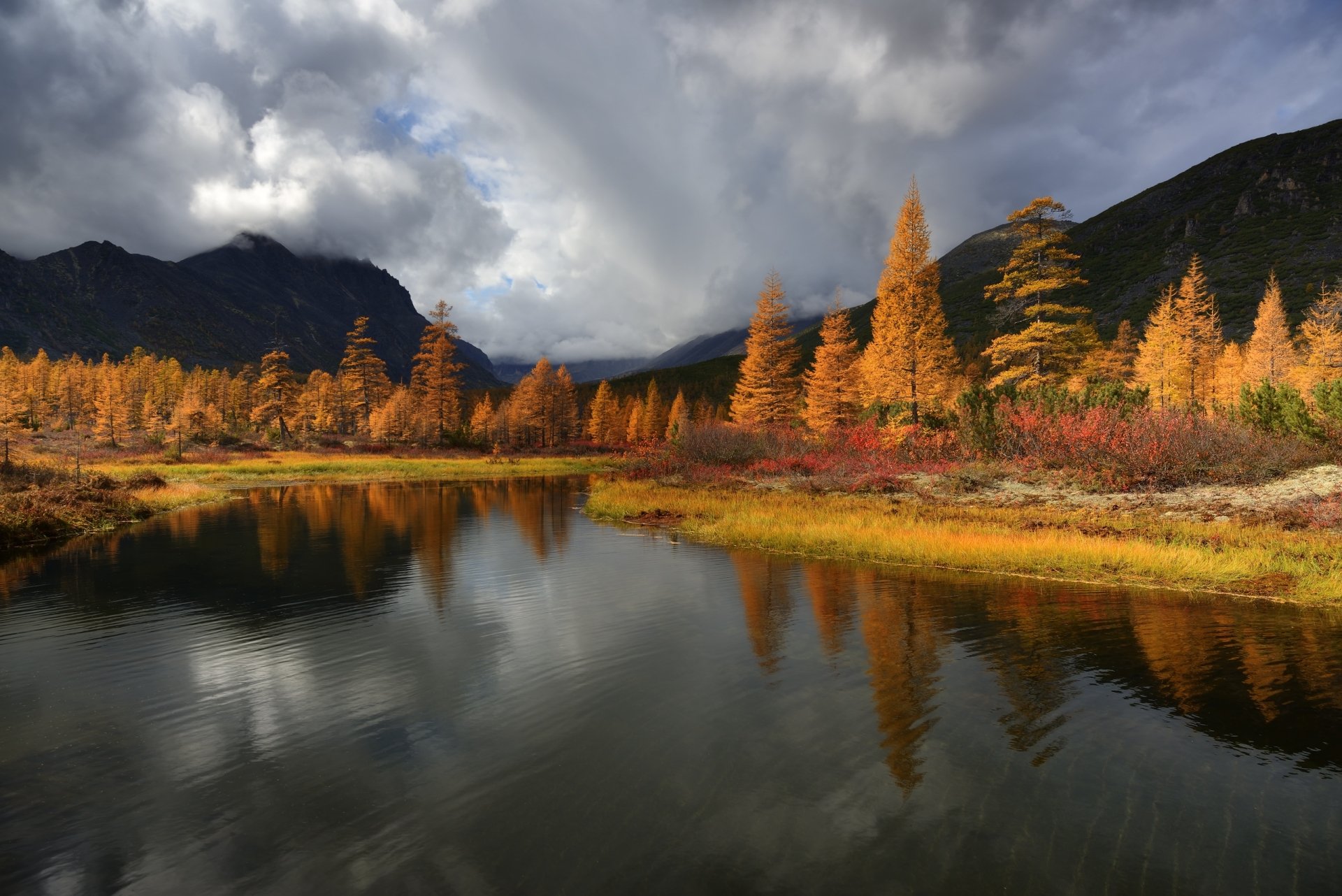 HD desktop wallpaper showcasing a serene fall landscape with vibrant autumn trees reflecting in a calm lake under a dramatic cloudy sky.