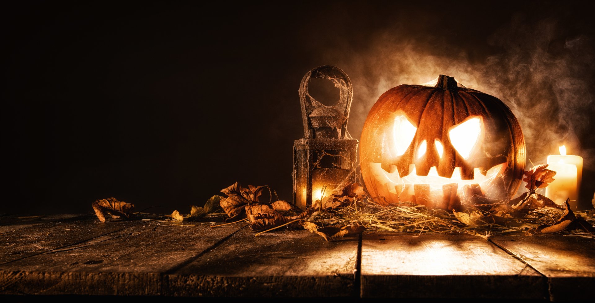 4K Ultra HD Halloween-themed PC desktop wallpaper featuring a glowing carved pumpkin, dried leaves, and a rustic lantern on a wooden surface with smoky background.
