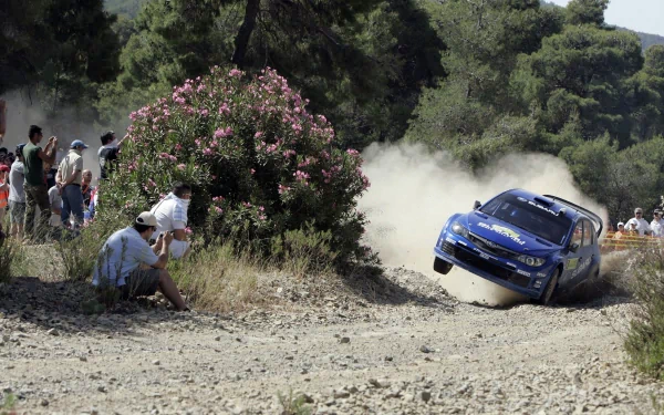 HD PC desktop wallpaper featuring a Subaru WRC racing car kicking up dust on a rugged dirt track, surrounded by spectators and lush greenery.