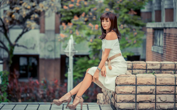 Elegant Asian woman with black hair in a white dress and high heels poses outdoors, captured with a soft depth of field in 4K Ultra HD quality.