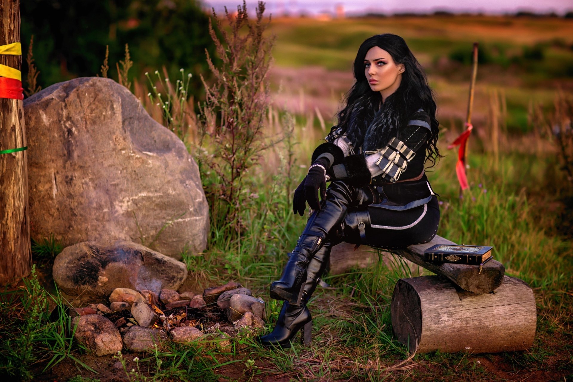 HD desktop wallpaper of a woman cosplaying Yennefer of Vengerberg from The Witcher 3: Wild Hunt, with long black hair and boots, seated outdoors near a campfire.