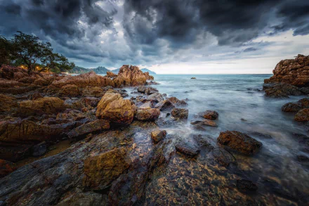 A stunning seascape in Thailand with a rocky coastline under dramatic, stormy clouds. The scenery showcases the natural beauty of the ocean and coast. This HD wallpaper captures the awe-inspiring nature.