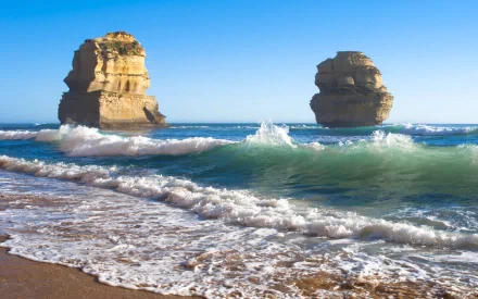 HD desktop wallpaper showing The Twelve Apostles rock formations rising from the ocean under a clear blue sky along a natural beach.