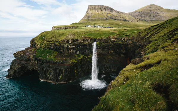4K Ultra HD view of Gásadalur waterfall cascading off a rugged cliff into the ocean on the Faroe Islands coast, framed by green hills under a cloudy sky.
