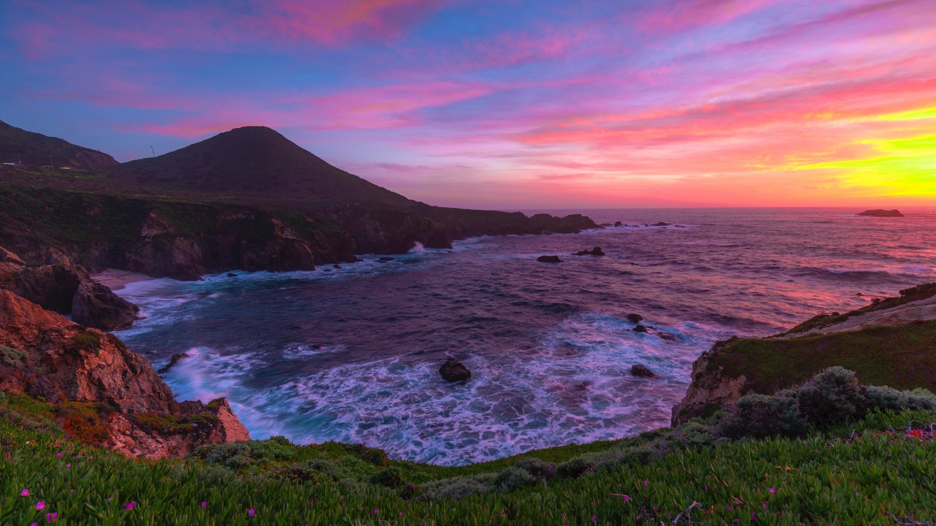 A vibrant 8K Ultra HD desktop wallpaper of a stunning coastal sunset, showcasing a colorful sky over the ocean with rugged cliffs and green foreground vegetation.