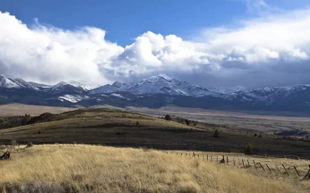 Rolling grassy hills and a wooden fence lead to snow-capped mountains under a blue sky with clouds in Malheur National Forest, John Day, Oregon.