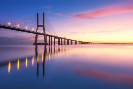 Vasco da Gama Bridge stretching across the Tagus River at dawn, pink and gold sky mirrored in calm water — a man-made span captured as an HD desktop wallpaper.