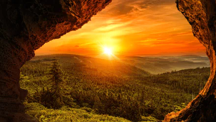 Sunbeam breaks over a lush forest at sunset, framed by the rocky opening of a cave in this vivid 4K Ultra HD nature landscape wallpaper.