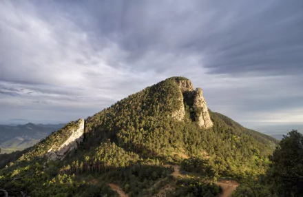 8K Ultra HD PC desktop wallpaper and background: forested hill rising beneath a dramatic cloudy sky, lush nature landscape with rocky outcrops.