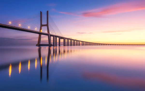 Vasco da Gama Bridge stretching across the Tagus River at dawn, pink and gold sky mirrored in calm water — a man-made span captured as an HD desktop wallpaper.