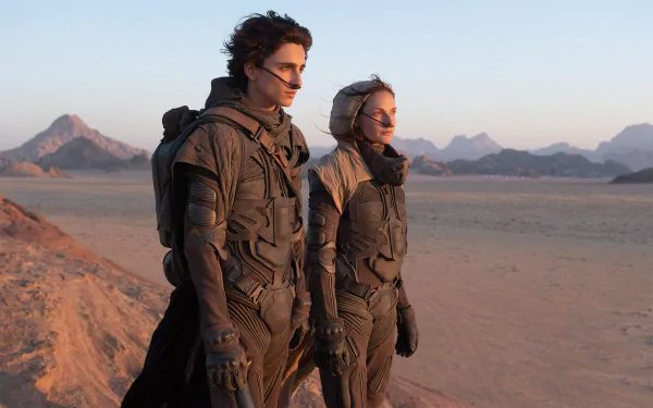 HD PC wallpaper featuring Rebecca Ferguson and Timothée Chalamet in desert gear from the movie Dune (2021), standing on a vast arid landscape with mountains in the background.