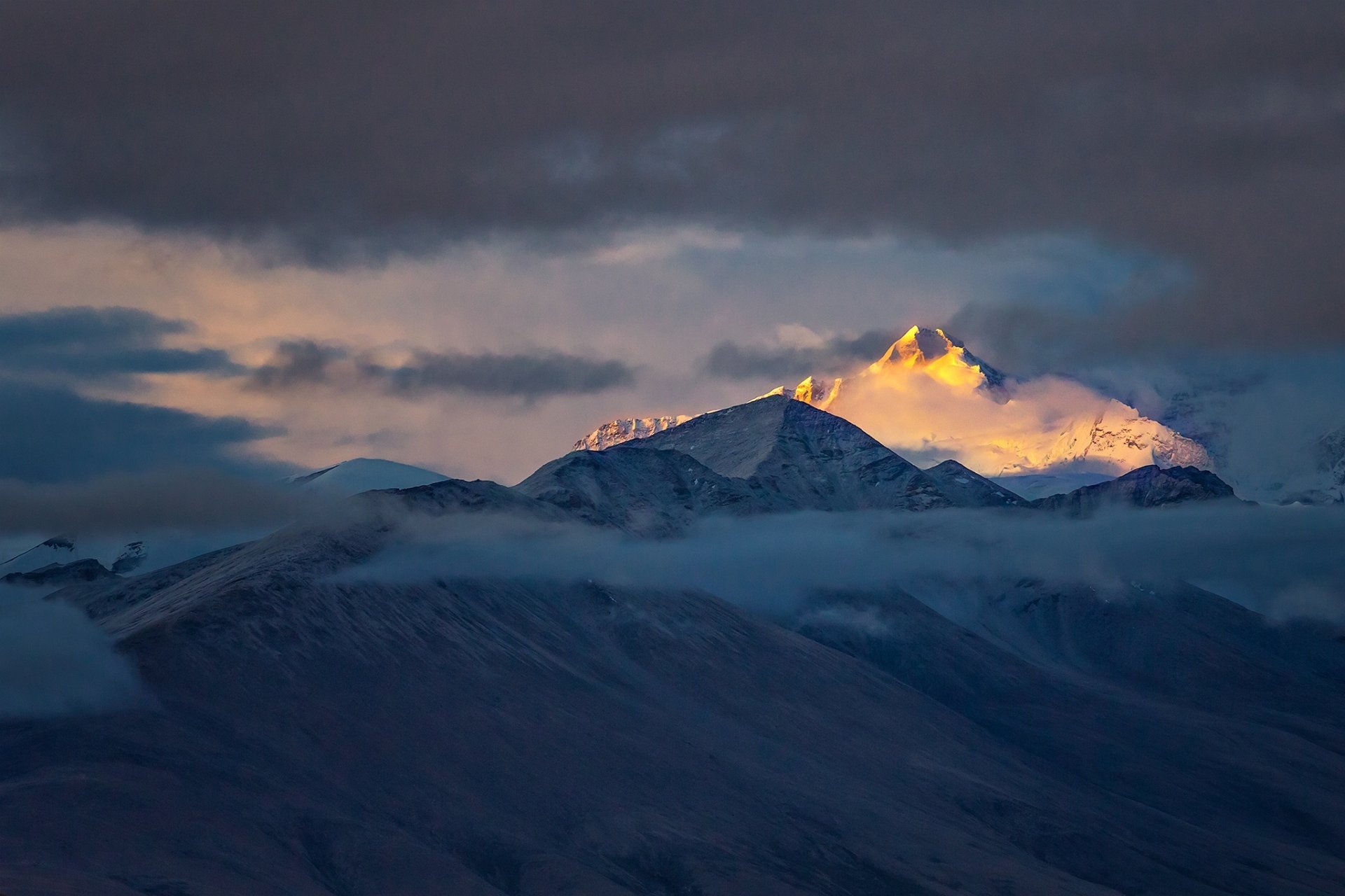 HD desktop wallpaper of a snow-capped mountain peak in Tibet, bathed in golden sunlight above a layer of clouds under a moody sky.