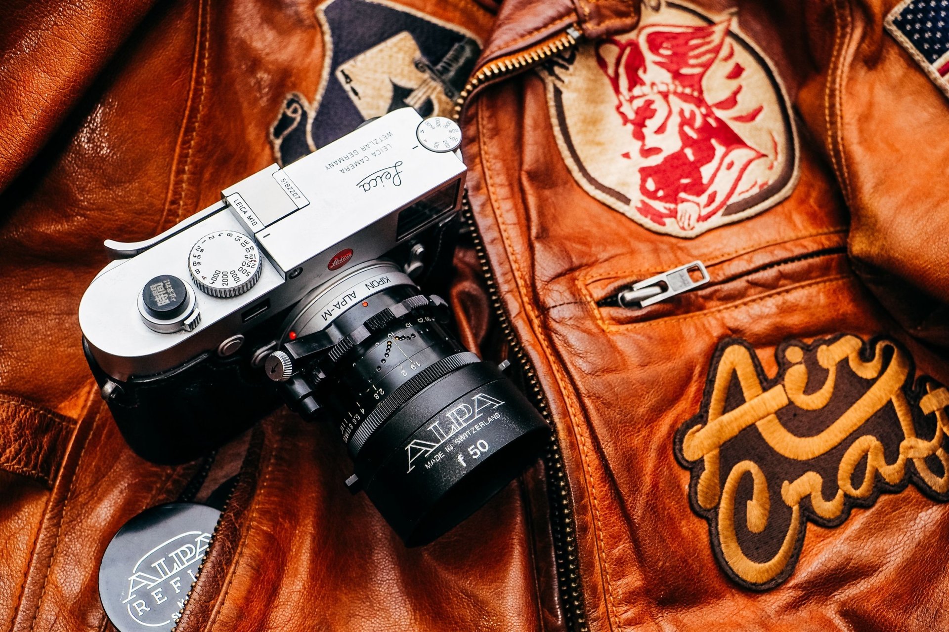 Man-made camera resting on a patched brown leather jacket, 2K Quad HD PC desktop wallpaper/background — close-up of a vintage film camera and textured leather.