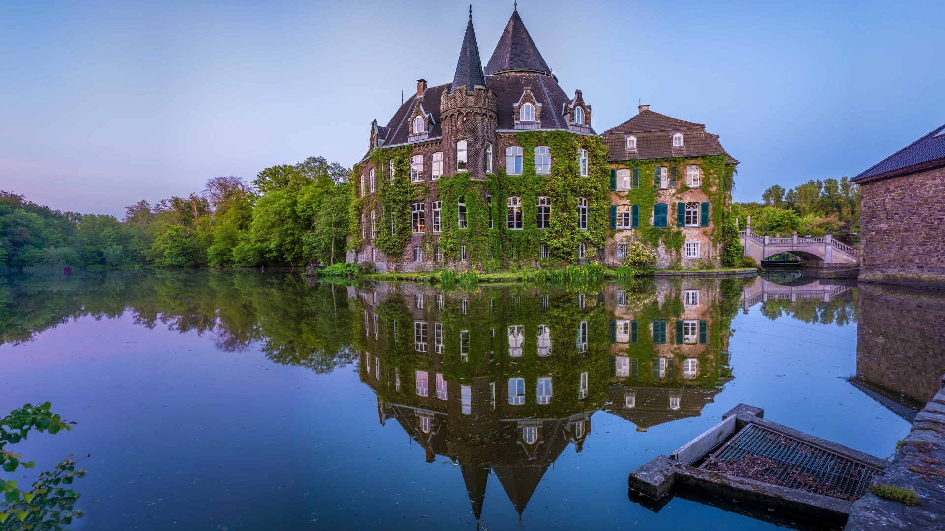 4K Ultra HD image of a man-made ivy-covered castle reflected in calm water, surrounded by lush greenery under a clear sky.