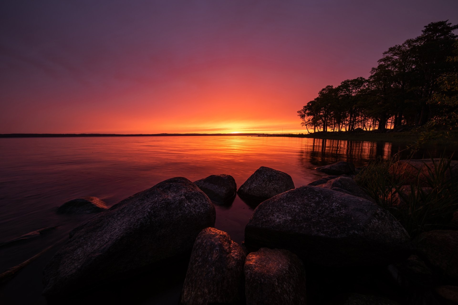 HD wallpaper of a serene Swedish lake at sunrise. The tranquil scene features a calm lake, large stones in the foreground, and a silhouetted tree line under a vibrant, colorful sky.
