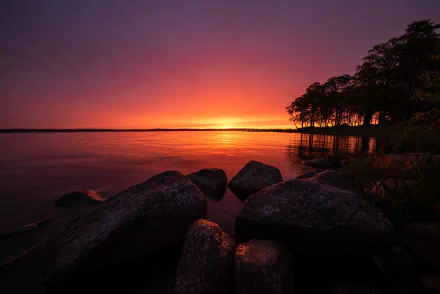 HD wallpaper of a serene Swedish lake at sunrise. The tranquil scene features a calm lake, large stones in the foreground, and a silhouetted tree line under a vibrant, colorful sky.