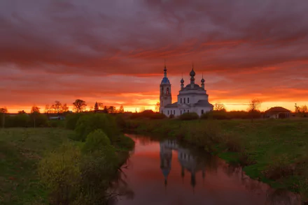 HD desktop wallpaper of a religious temple and church by a river at sunset, with vibrant orange and red skies reflecting on the water.