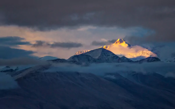 HD desktop wallpaper of a snow-capped mountain peak in Tibet, bathed in golden sunlight above a layer of clouds under a moody sky.