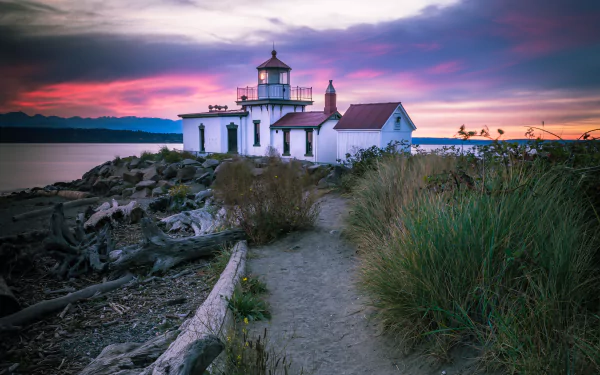 5K Ultra HD PC desktop wallpaper of a man-made lighthouse on a rocky shore at sunset, grasses and a sandy path leading to a white keeper’s house beneath a purple-pink sky.