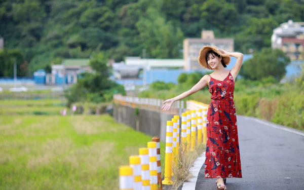 A woman in a red floral dress and wide-brimmed hat poses confidently on a rural road, captured in vibrant 4K Ultra HD quality.