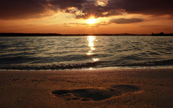 HD desktop wallpaper of a serene beach shore at sunset with golden sunlight reflecting on the water and a footprint imprinted in the sand.