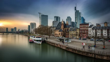 A 4K Ultra HD view of Frankfurt, Germany, featuring a riverfront with historic and modern buildings along the water under a calm, overcast sky.