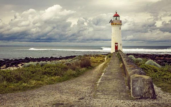 4K Ultra HD desktop wallpaper of a man-made lighthouse standing at the edge of a rocky shore under a cloudy sky.