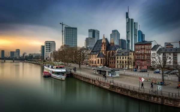 A 4K Ultra HD view of Frankfurt, Germany, featuring a riverfront with historic and modern buildings along the water under a calm, overcast sky.